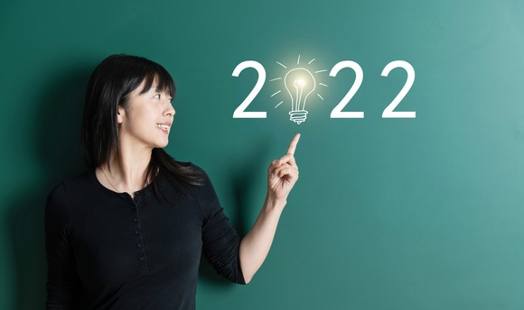A smiling person pointing to 2022 written on a chalkboard with a lightbulb in the place of the 0.