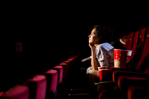 A woman sitting in a movie theater in a row of empty seats, looking up toward the screen, with popcorn and a soft drink ready to hand.