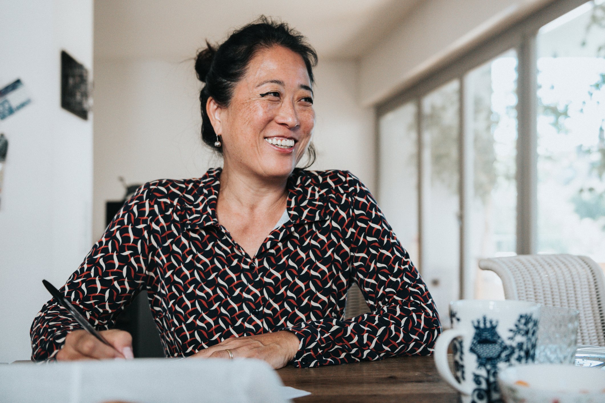 A lady smiling while sitting at a wooden desk with a pen in her right hand.