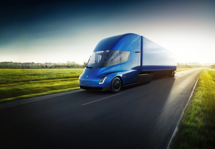 Blue Tesla Semi driving down an empty road surrounded by grass.