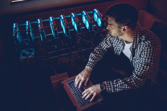 A person sitting next to a mining rig using a laptop to mine Bitcoin.