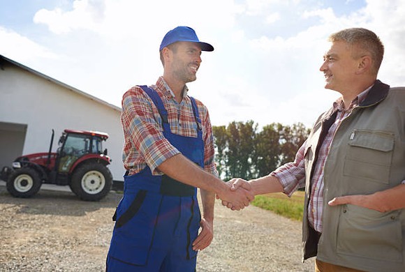 Two men shaking hands on a wide parking space on what looks like a farm.