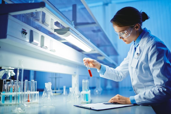 A scientist at a laboratory bay looks at a test tube while resting a hand on a clipboard with a pen.