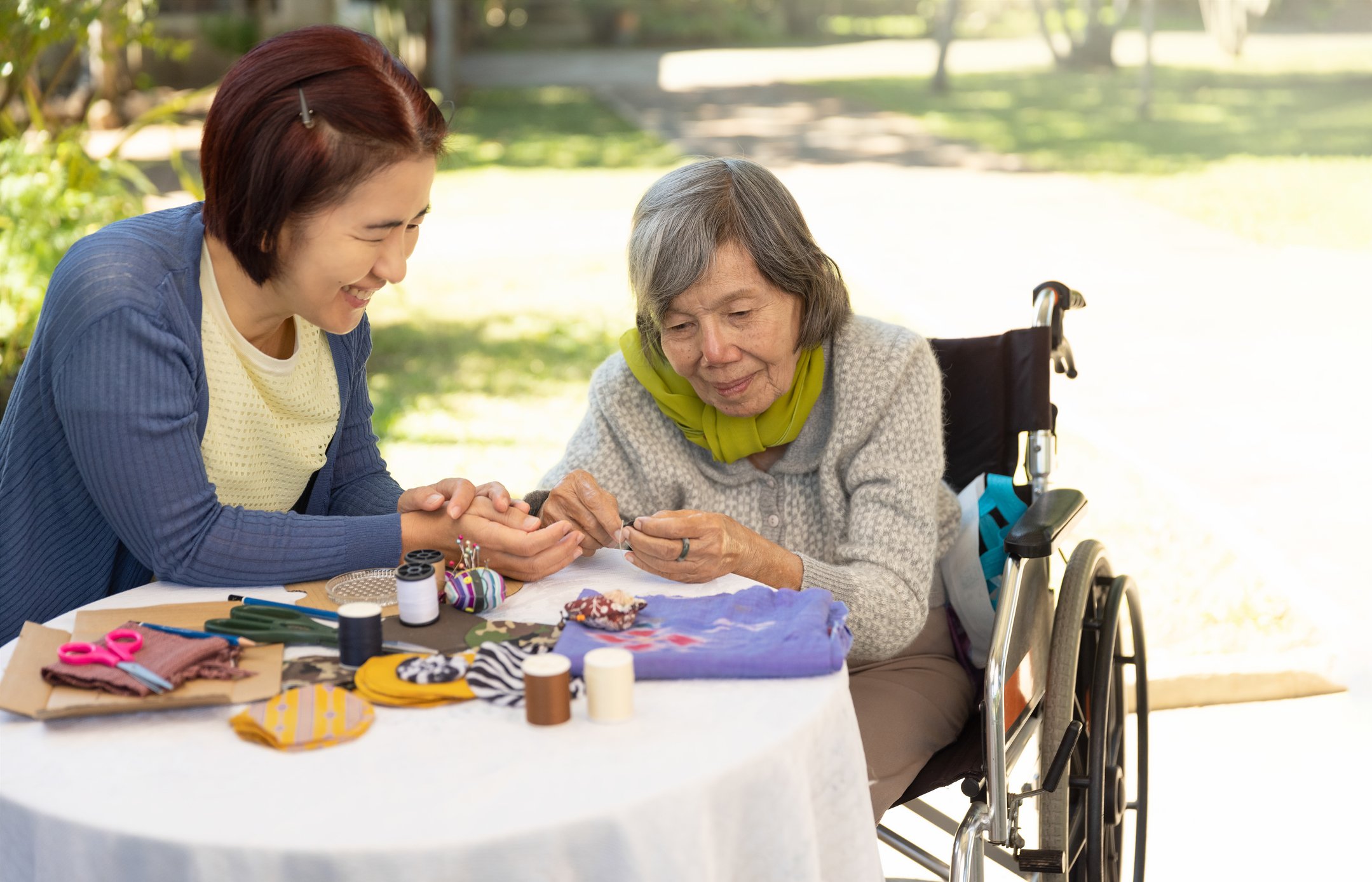 A caretaker helps an older woman with needlepoint.