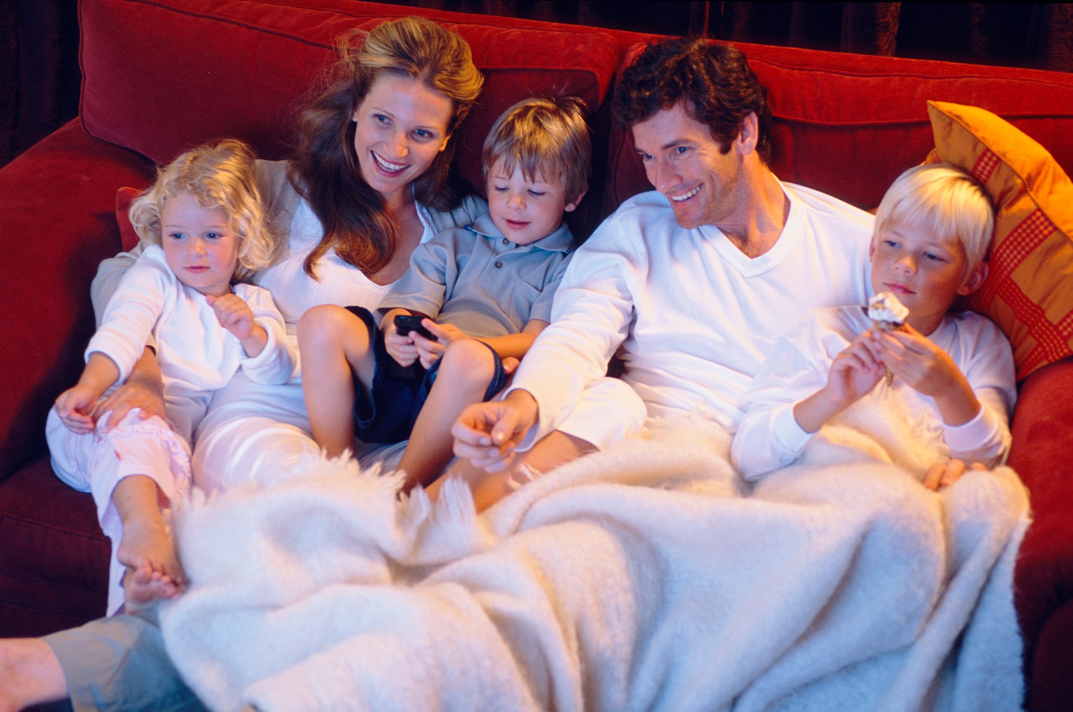 A young couple with three children all huddled on the couch watching television in a darkened room.