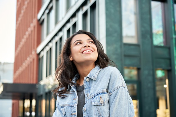 A young woman in a denim jacket smiles as she stares up into the sky in a city setting. 