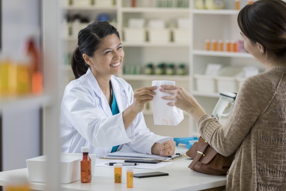 A smiling pharmacist hands a bagged purchase to a customer. 