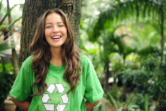 Smiling person wearing a T-shirt with a recycling symbol leans against a tree outside.