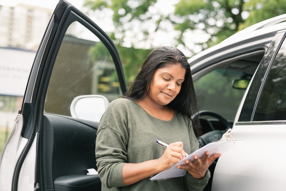 A person standing next to a car writes on a clipboard.