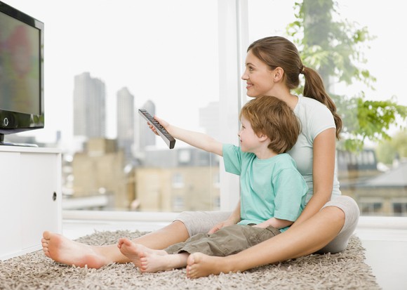 A young parent and child sitting on a throw rug watching television together.