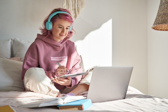 Student sitting in bed in front of a laptop and taking notes.