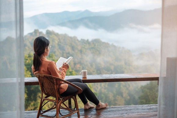 A person reading a book on balcony with coffee at her side.