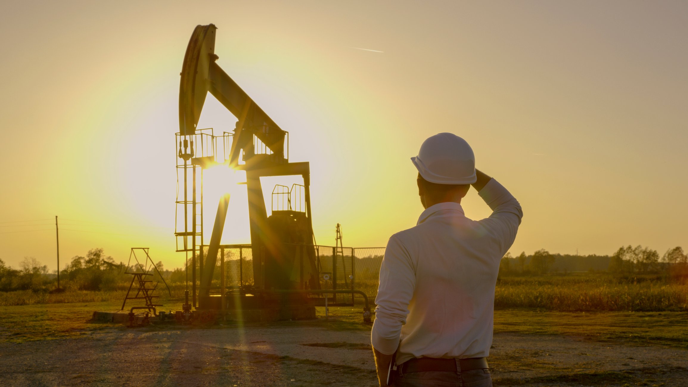 A person wearing a white hard hat looks at a pumpjack in front of a bright full yellow sun.