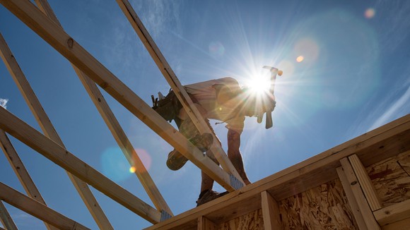 Person wielding a hammer on top of the frame of a house with the sun in the background.