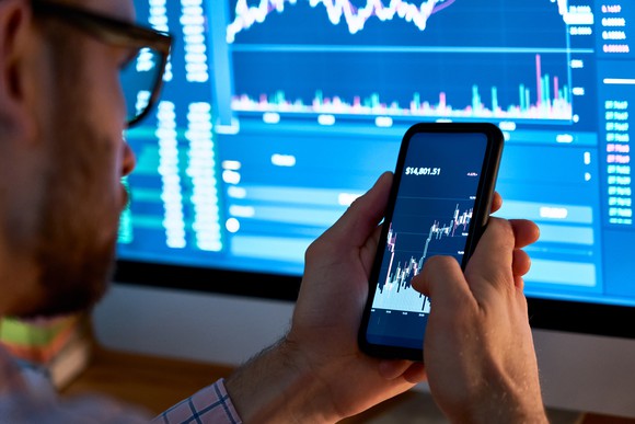 Man holding phone in front of computer screen showing market data.