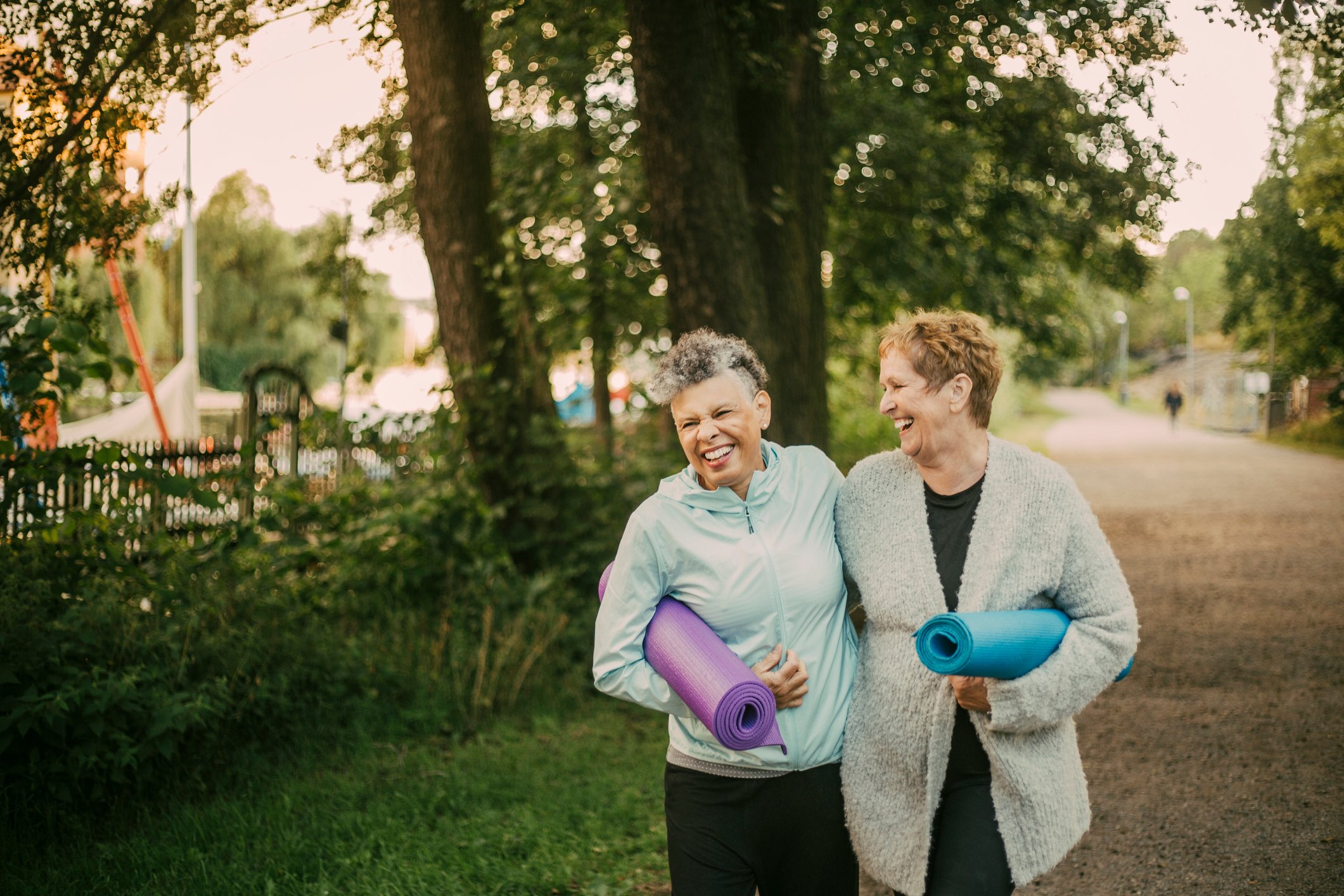 Seniors laugh while walking outside in the woods holding yoga mats.