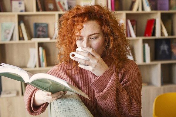 A person reads a book on a sofa while drinking a cup of coffee.