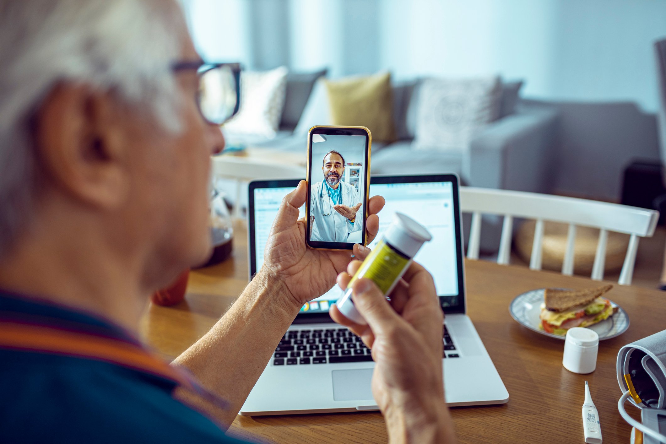 A person with silver hair holds a prescription bottle up to the camera during a telehealth interaction with a healthcare provider.