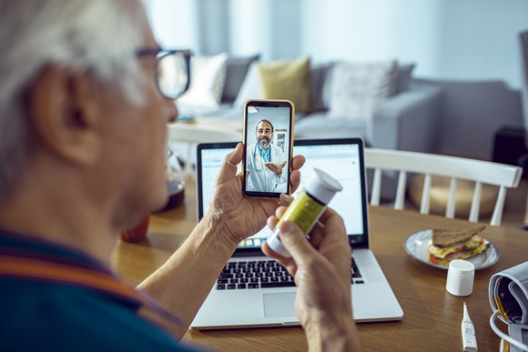 A person with silver hair holds a prescription bottle up to the camera during a telehealth interaction with a healthcare provider.