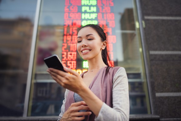 A woman stands in front of a financial information panel outside in a city and looks at something on her phone.