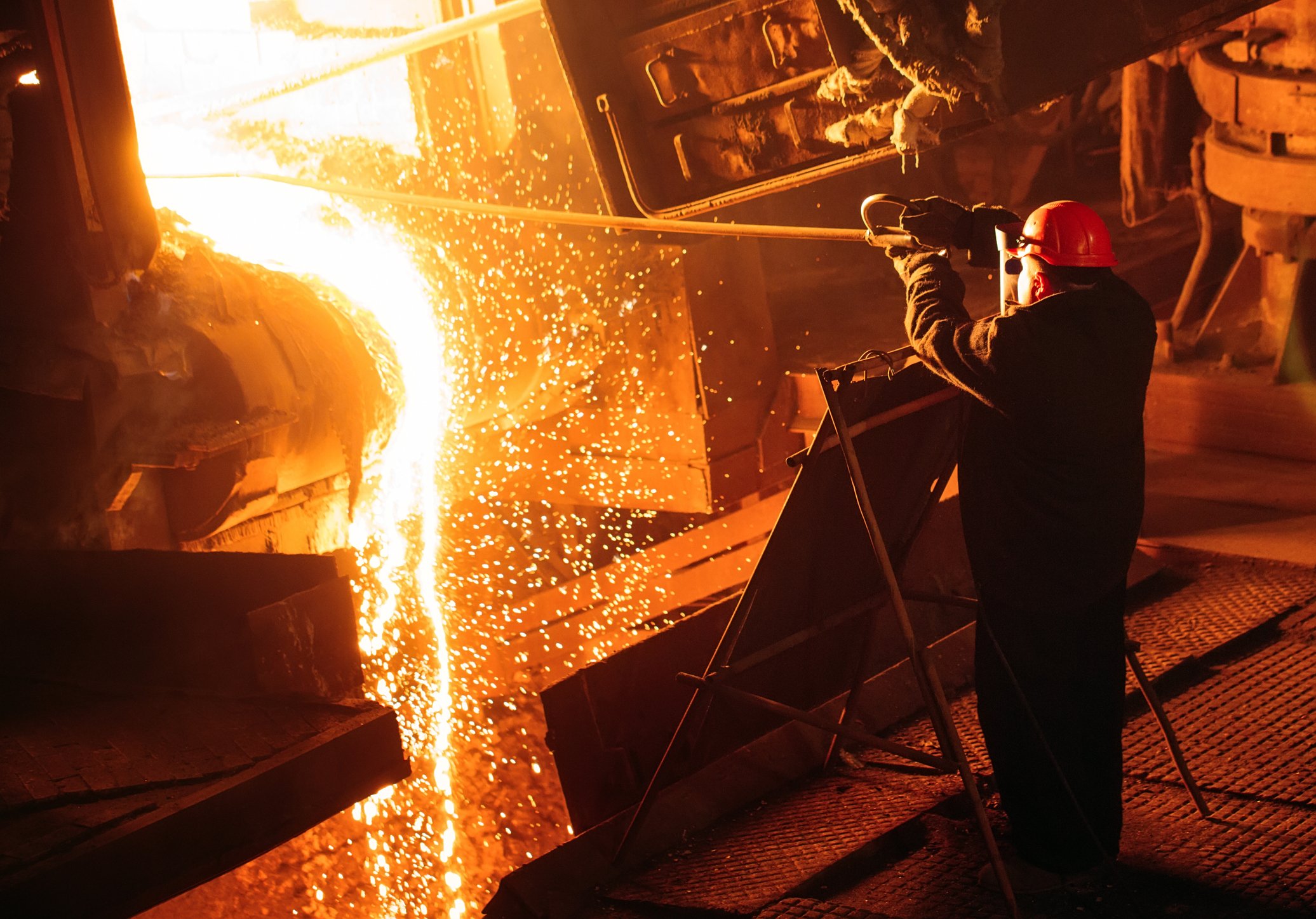 A steelworker tends a fiery furnace in protective gear. 