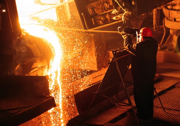 A steelworker tends a fiery furnace in protective gear. 
