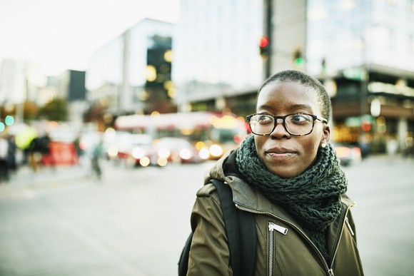 Confident adult wearing scarf and coat smiles while walking on street outside.