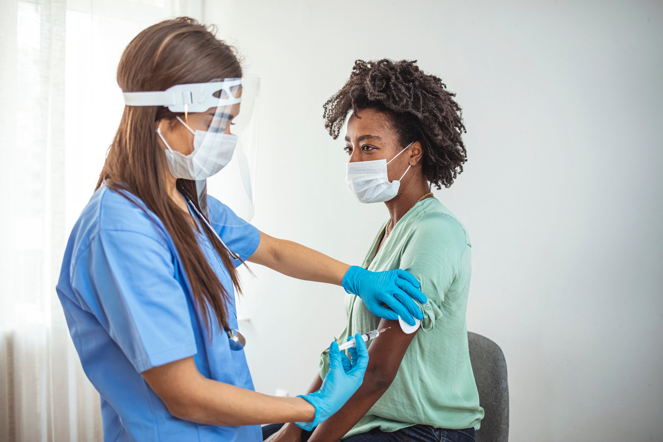 A caregiver in personal protective equipment administers a shot to a patient wearing a mask.