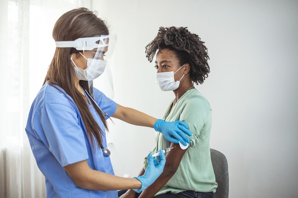A caregiver in personal protective equipment administers a shot to a patient wearing a mask.
