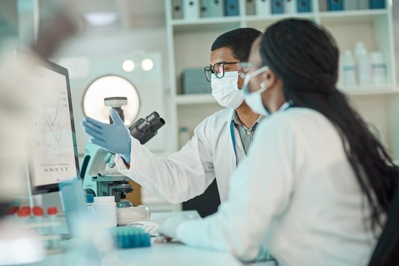 Two face-masked medical researchers having a discussion at a laboratory table.