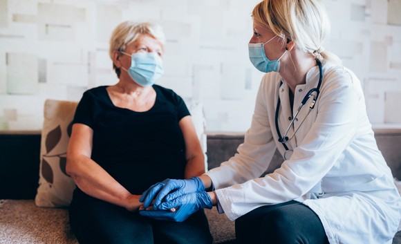 A healthcare worker in surgical mask and gloves holds the hands of a patient, also wearing a surgical mask.