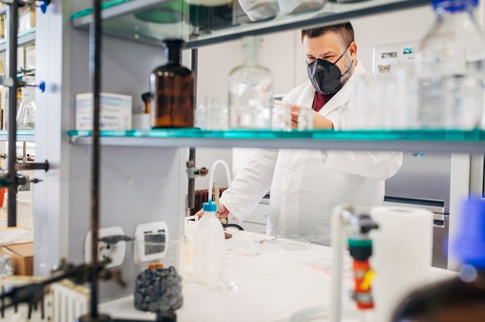 A researcher examines a test tube while standing at a cluttered laboratory bench.