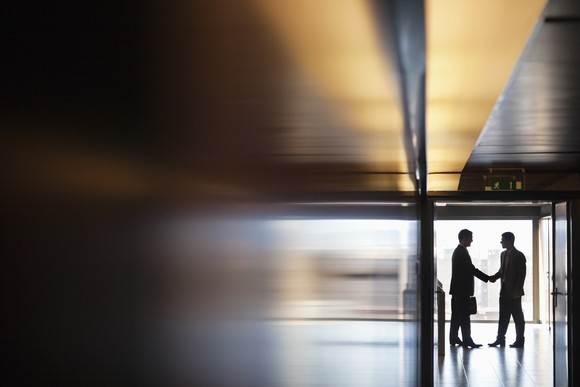 Two executives shake hands at the end of a long hallway. 