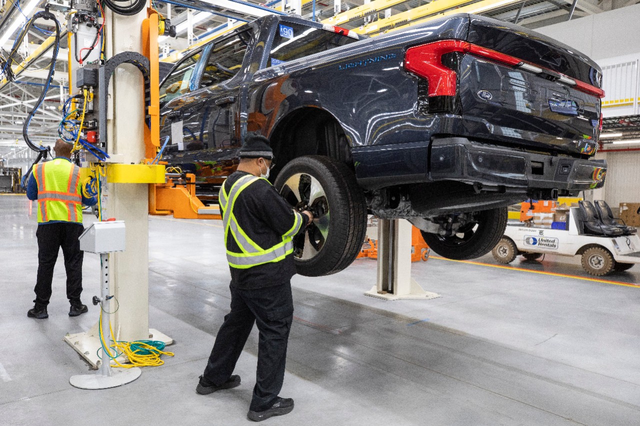 Assembly line workers install tires on an all-electric Ford F-150 Lightning vehicle that has been raised off the ground