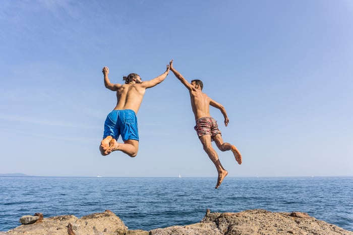 Two people by the sea are leaping into the water while high-fiving each other.