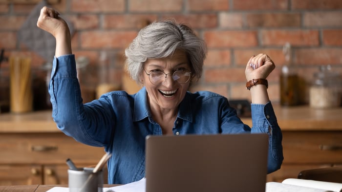 An older person smiling with arms raised while looking at a laptop. 