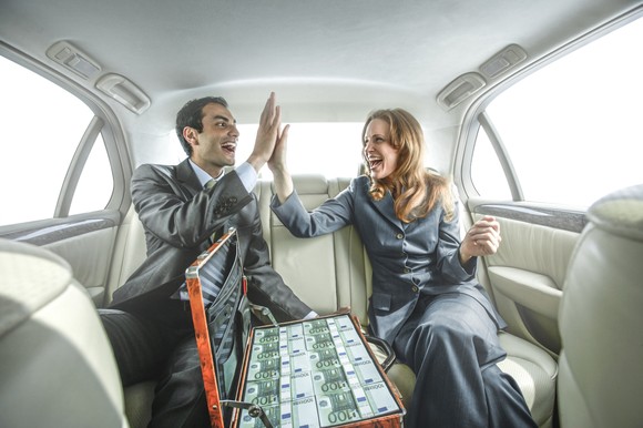 Two people sitting in the back seat of a car smile and high-five each other over a suitcase full of money.