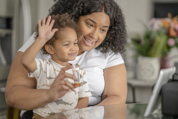 An adult holds a child and a bottle of pills in front of a tablet as part of a medical visit.
