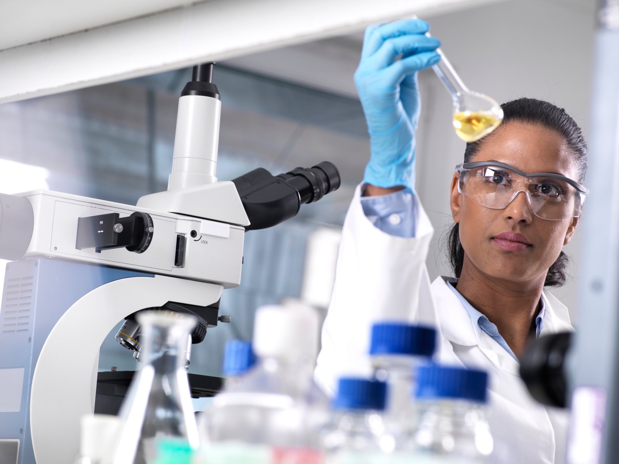 A lab researcher wearing safety goggles and rubber gloves looks at a liquid in a lab flask.