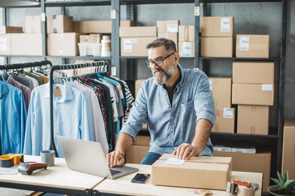 Person typing on a laptop and labeling a shipping box, with racks of clothing and boxes in the background.