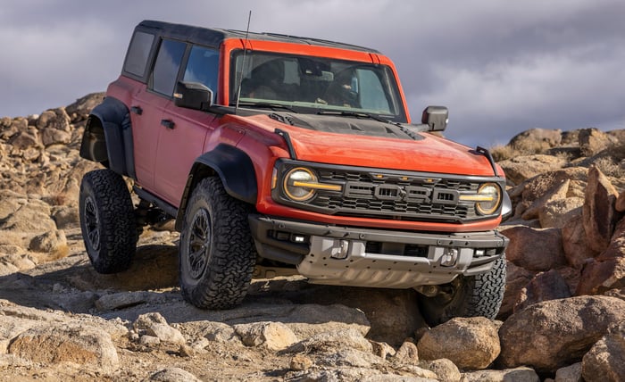 An orange 2022 Ford Bronco Raptor, an aggressive high-performance off-road SUV, shown traversing a rocky surface.