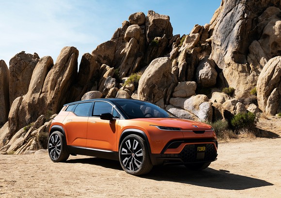 An orange Fisker Ocean, an electric SUV, parked on a rocky beach. 