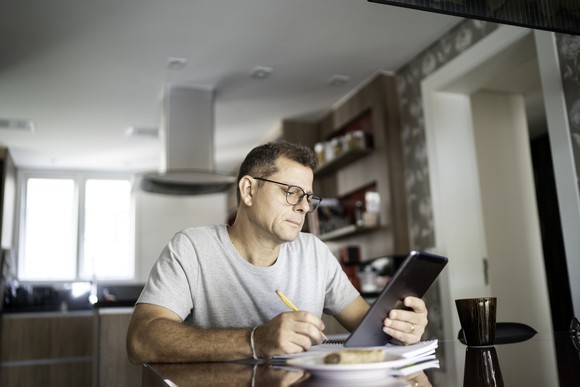 An investor sitting at the kitchen table and taking notes while using a tablet computer.