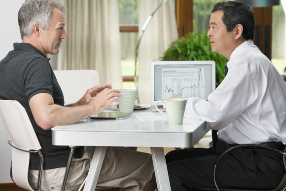 Two investors talk while sitting at a table in front of a laptop and two cups of coffee.