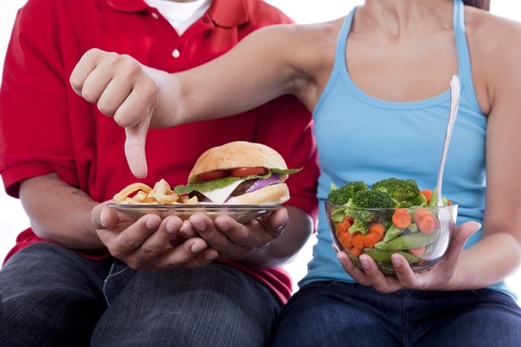 Torsos of two people with food items, with the woman showing a thumbs down above a plate with a burger.
