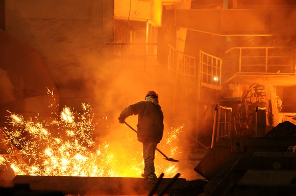 A worker holding a shovel in a factory with splashing molten iron in the background. 