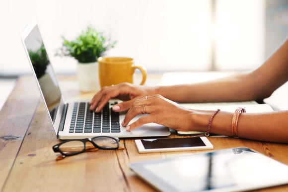 Hands typing on a laptop with glasses and mug on desk nearby.