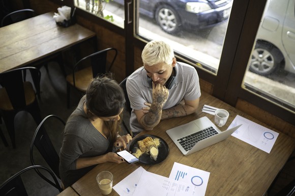 People working at restaurant table with food, paperwork, and a laptop.