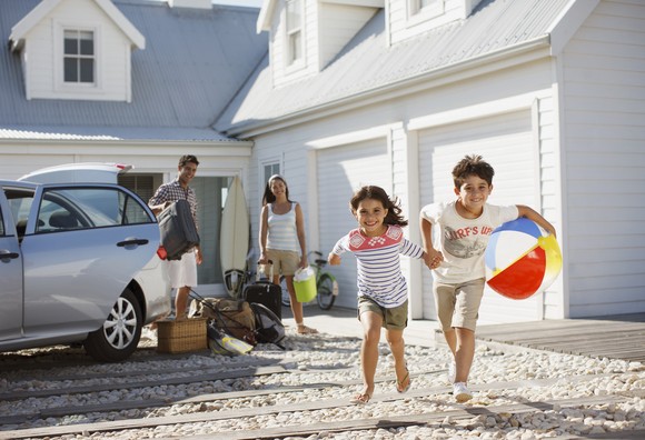 Two children hold hands and run, with one holding a beach ball. Two adults stand by a car with suitcases.