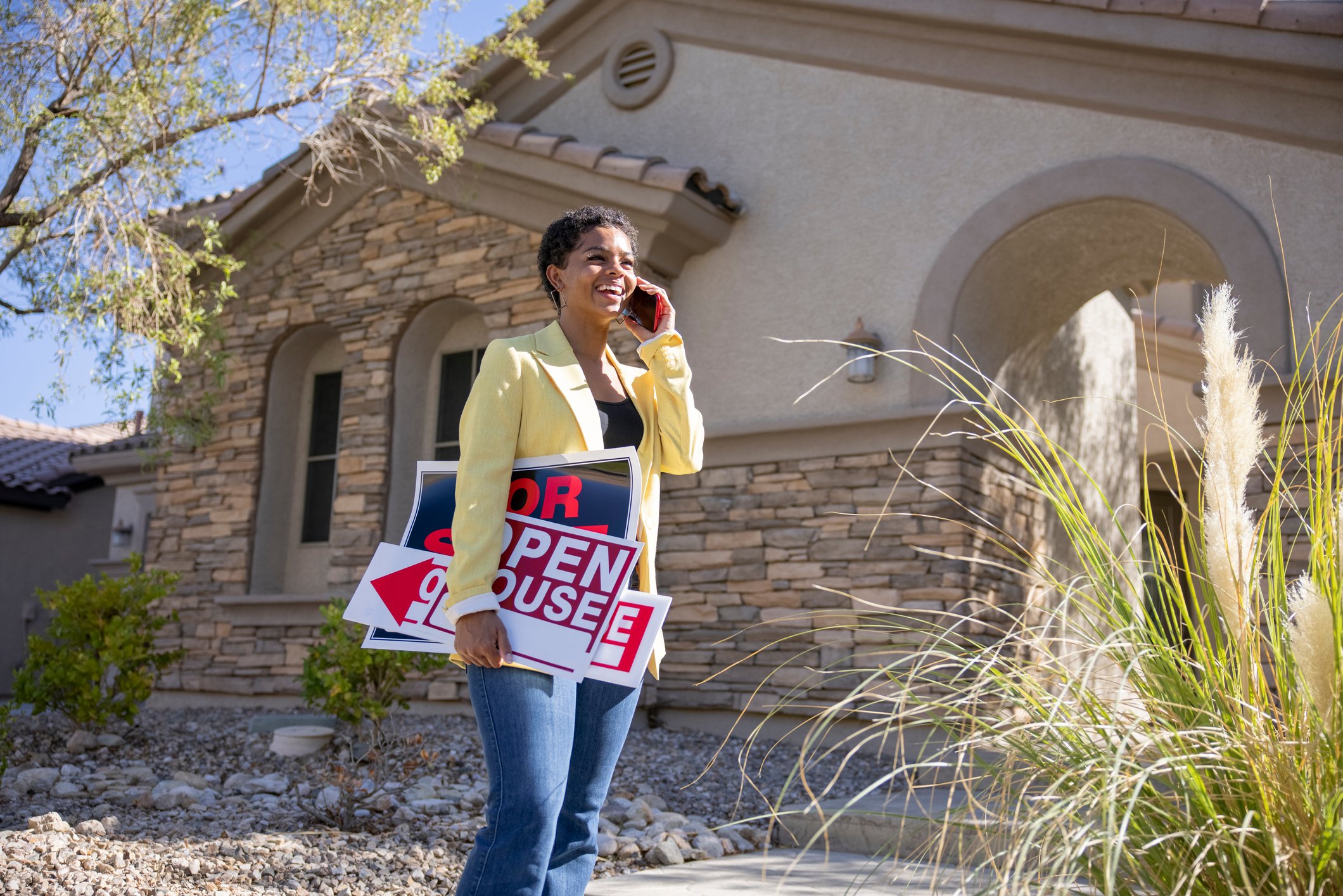 Person wearing a yellow jacket, standing outside of a house on a cell phone, holding a open house and for sale sign.